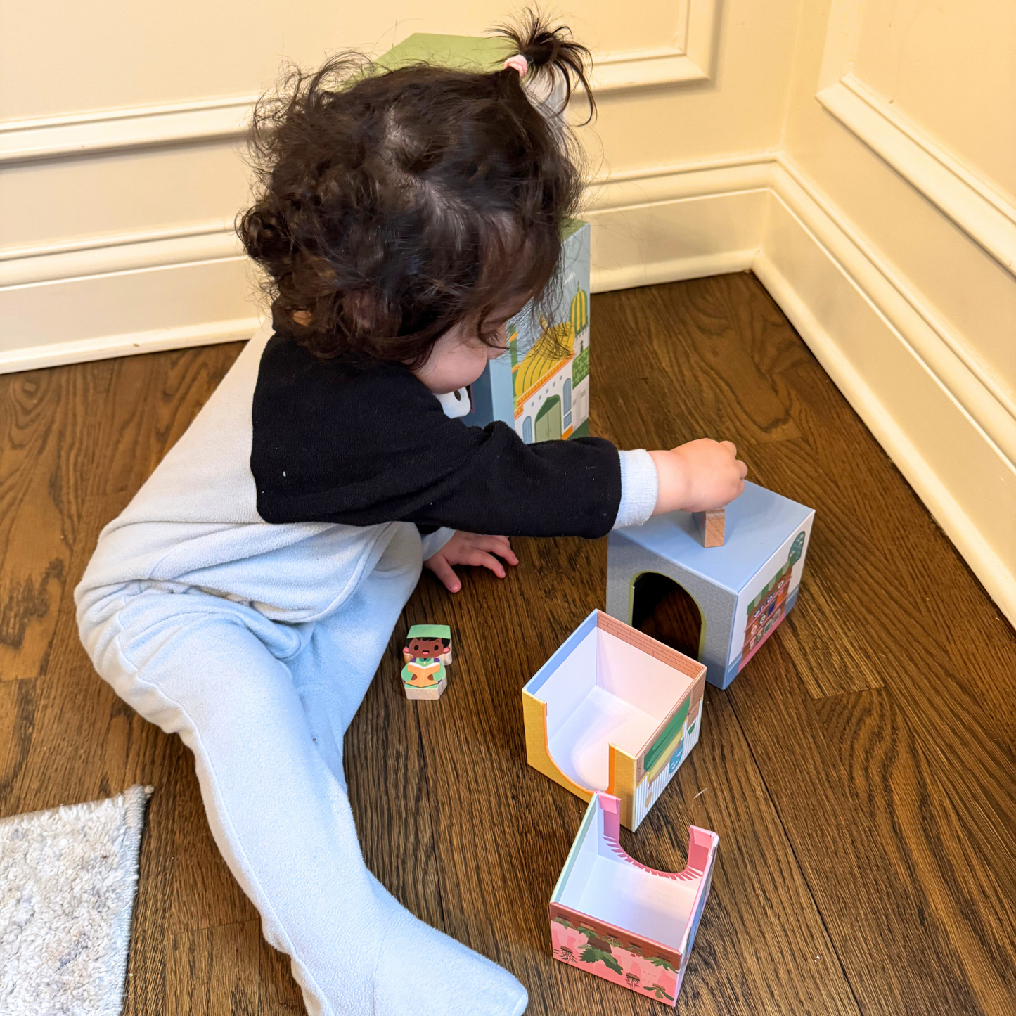 Toddler playing with stacking houses and toy figures on wood floor. Toy houses include masjid, home, school, garden and store.