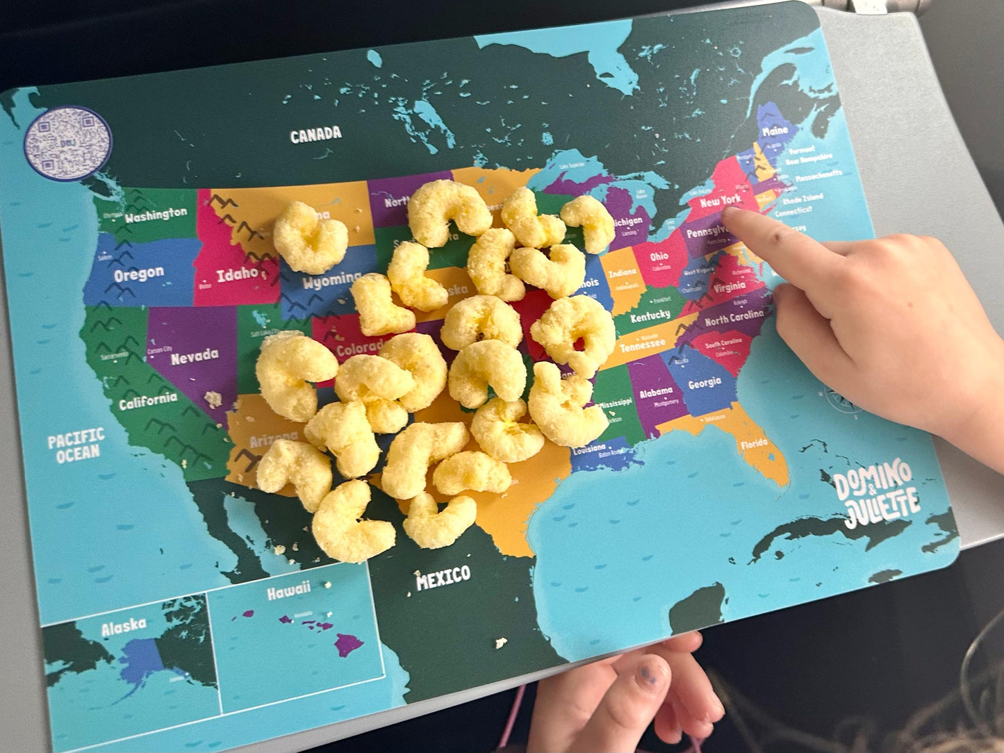 USA Map Placemat with Snack Puffs & Child's Hand Pointing to New York State. Domino & Juliette Brand.
